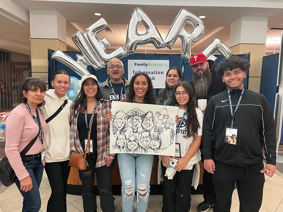 A group of people stands together in a shopping area holding a caricature drawing, with balloons spelling "WE ARE" in the background.