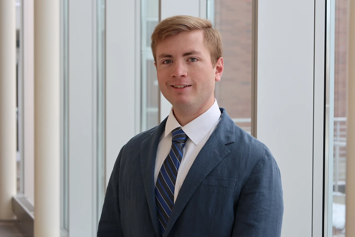 A man in a suit and tie is standing in front of large windows, smiling at the camera.