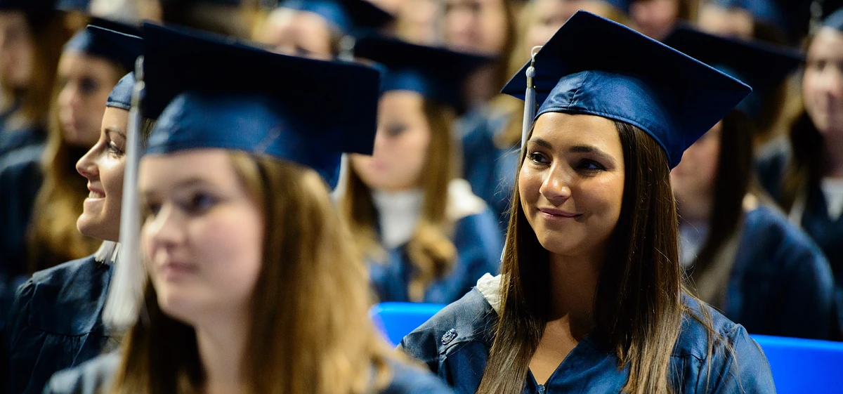 A group of people in blue graduation caps and gowns are sitting together, with one person smiling prominently.