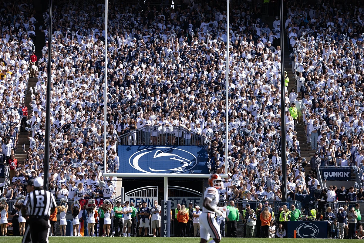 A large crowd of enthusiastic fans, many dressed in blue and white, fill the stands at a football game, watched by players and officials on the field.