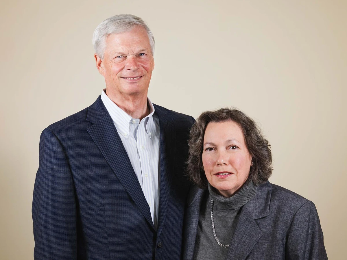 An older man and woman, dressed in formal attire, stand side by side against a neutral background.
