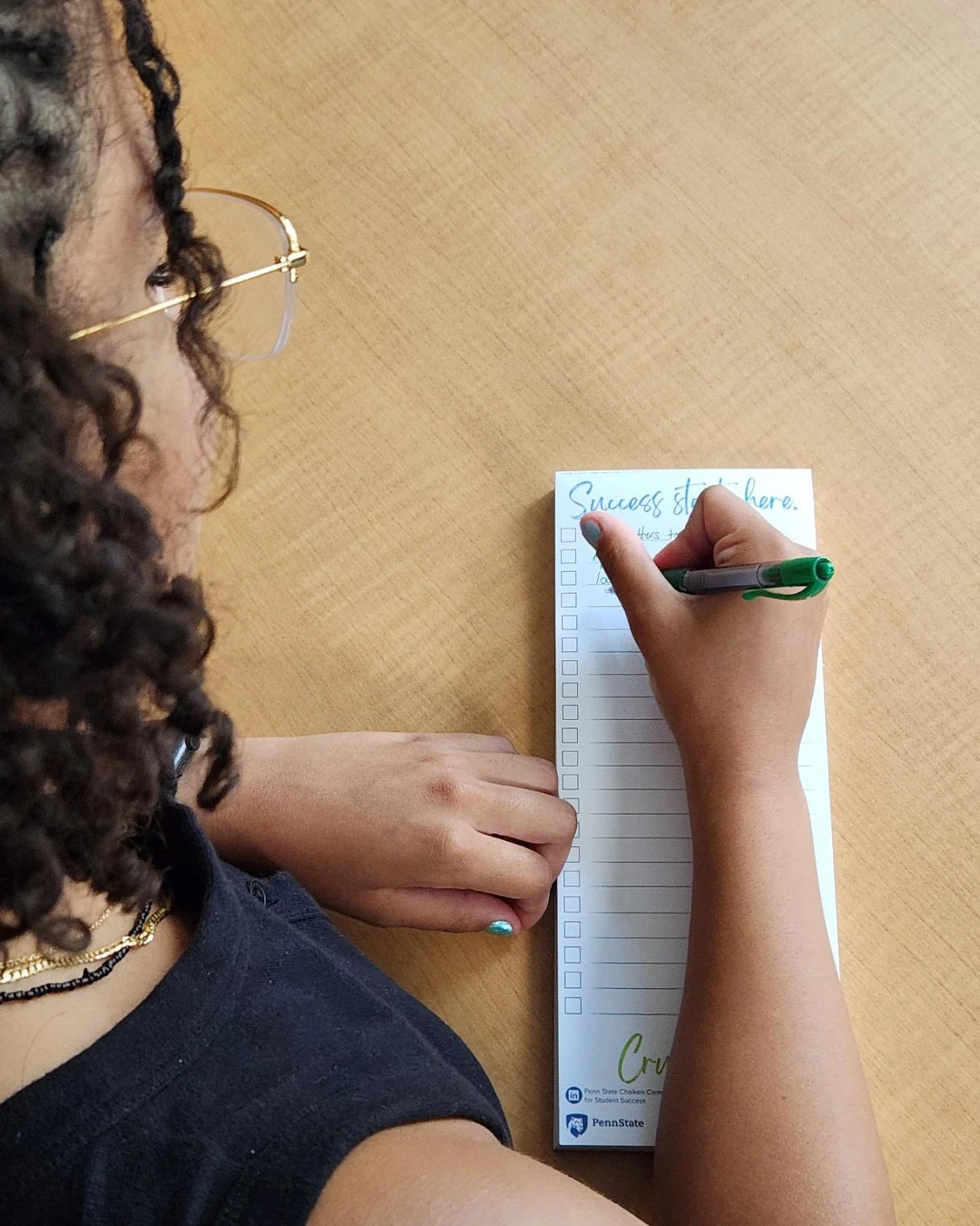 A person with curly hair and glasses writes on a notepad with checkboxes at a wooden desk.