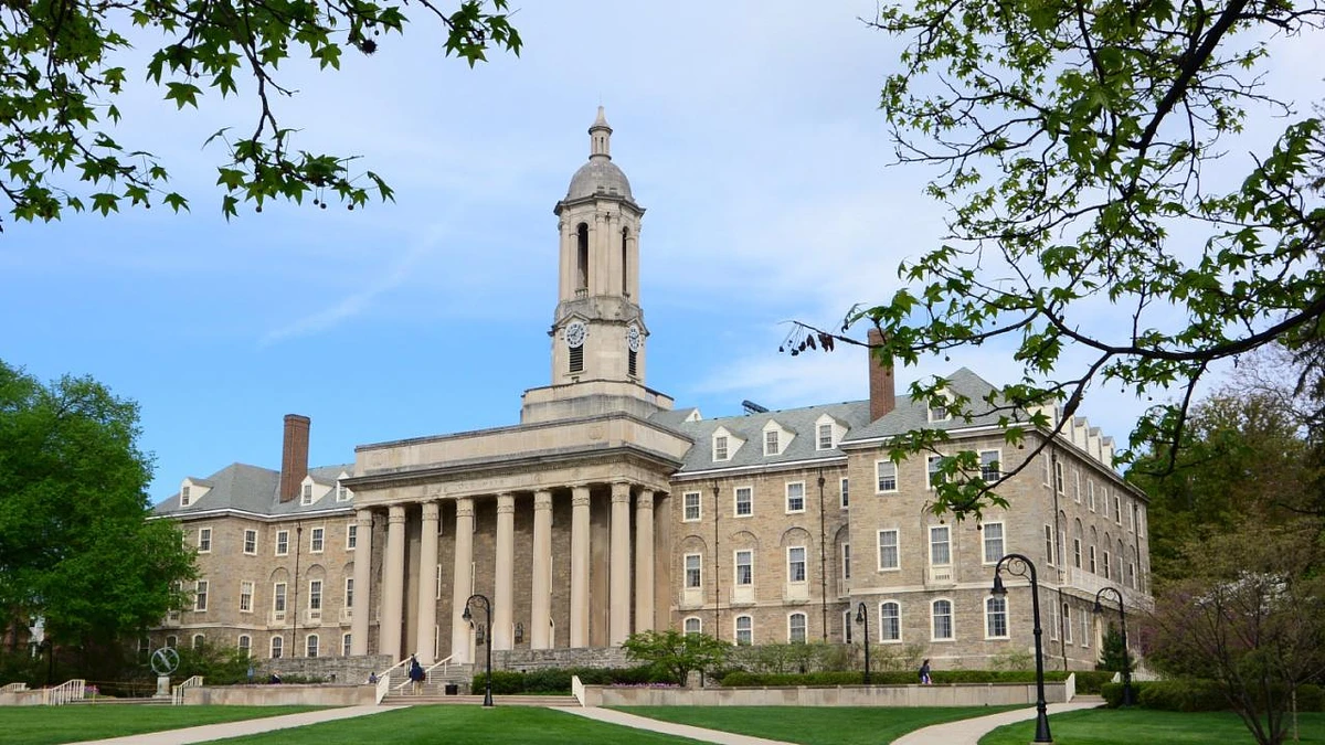 A large, historic building with a clock tower and columns is surrounded by trees under a clear blue sky.